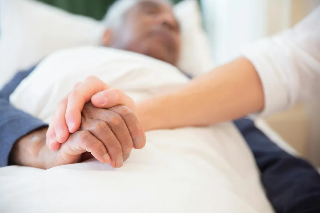 Close up of a nurse holding a seniors hand as he lays in bed