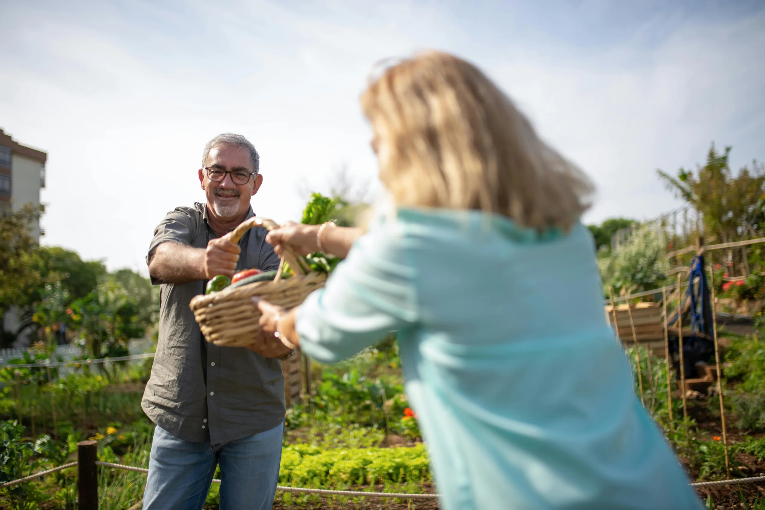 A lady hands a basket filled with vegetables to an elderly man with glasses