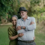 An senior woman stands behind an elderly man with her arms around him looking content