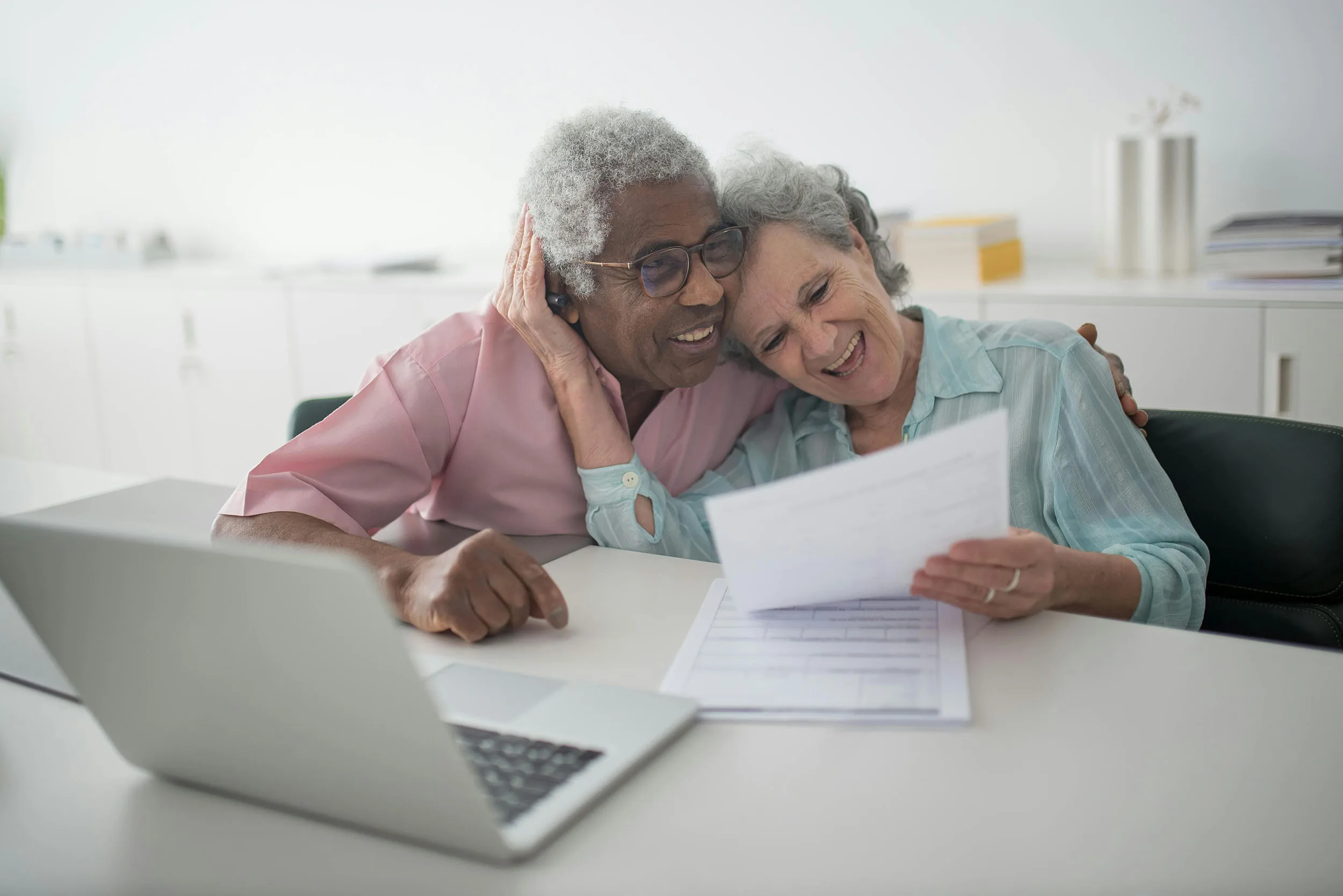 Two seniors at a computer laugh and hug as they look at papers