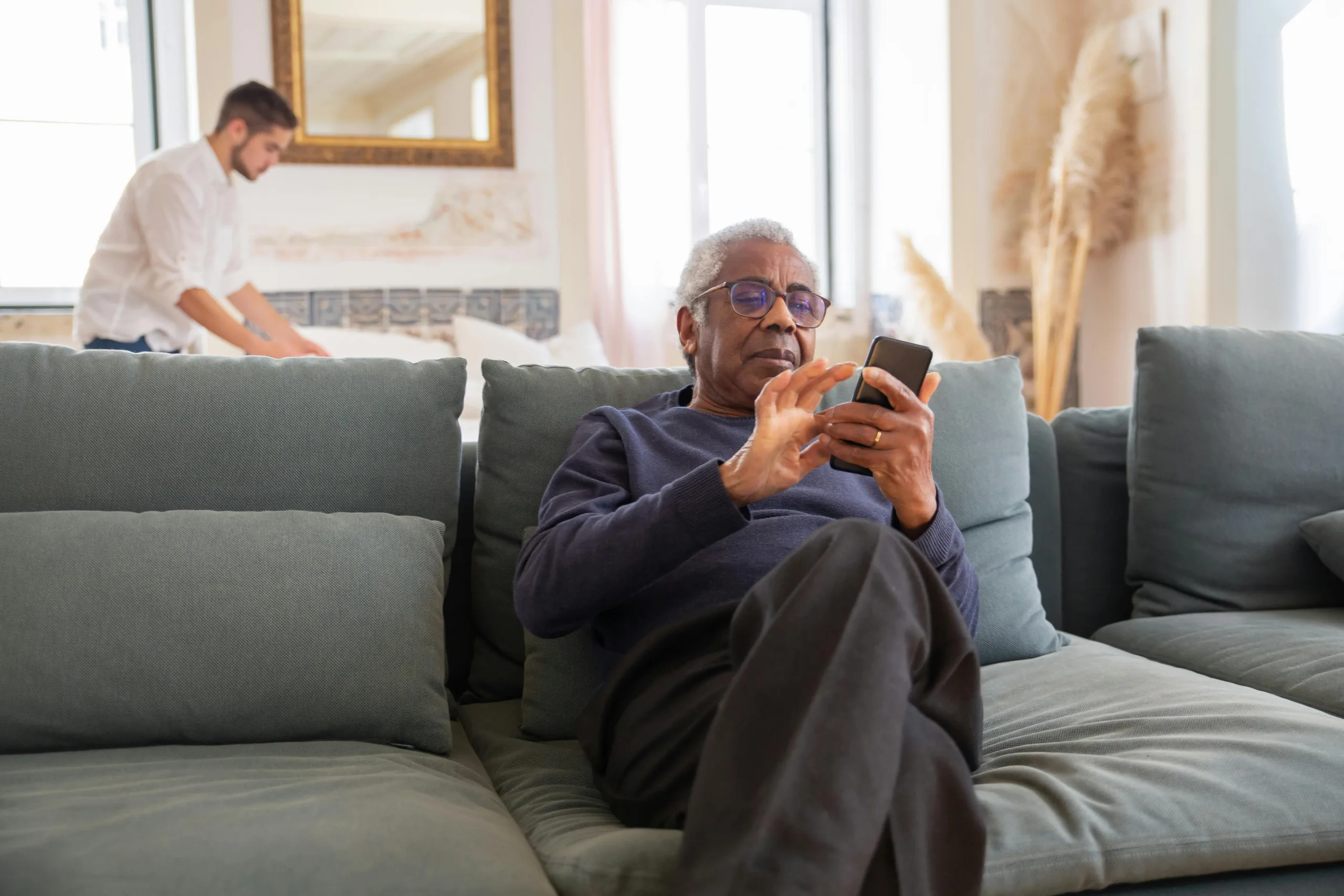 An elderly man on his phone sitting on the couch while a man makes his bed in the background