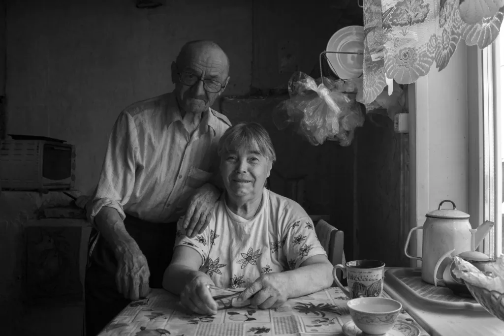 An elderly man with glasses stands next to a woman as she sits in the kitchen