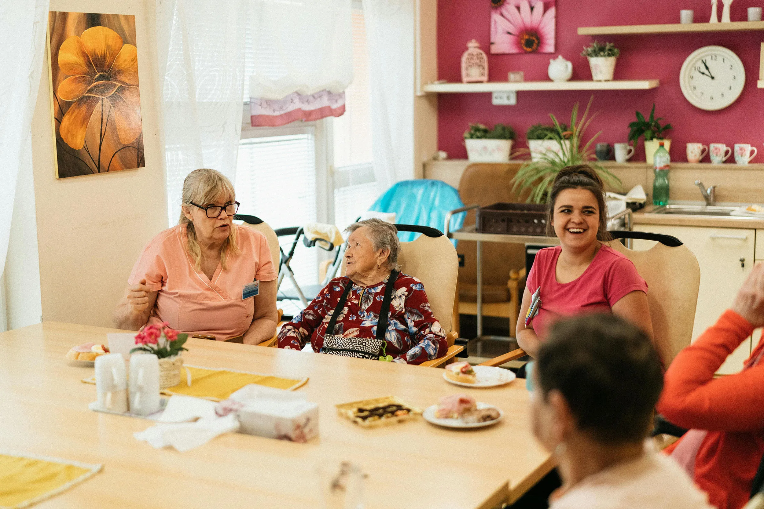 Three elderly woman sitting at a table with two staff members talking and laughing with them 