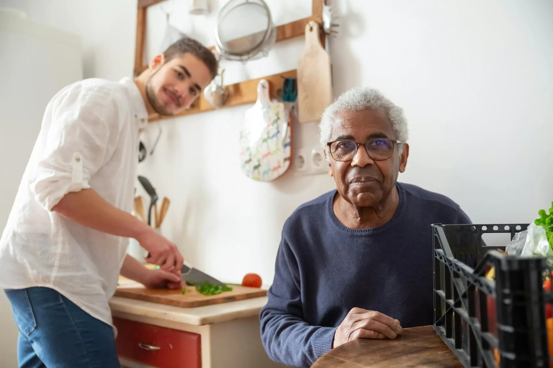 An elderly man with glasses smiles while another man prepares food