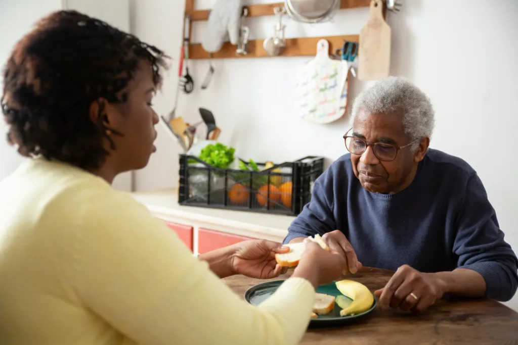 An elderly man sits at a table with a woman as she helps prepare food