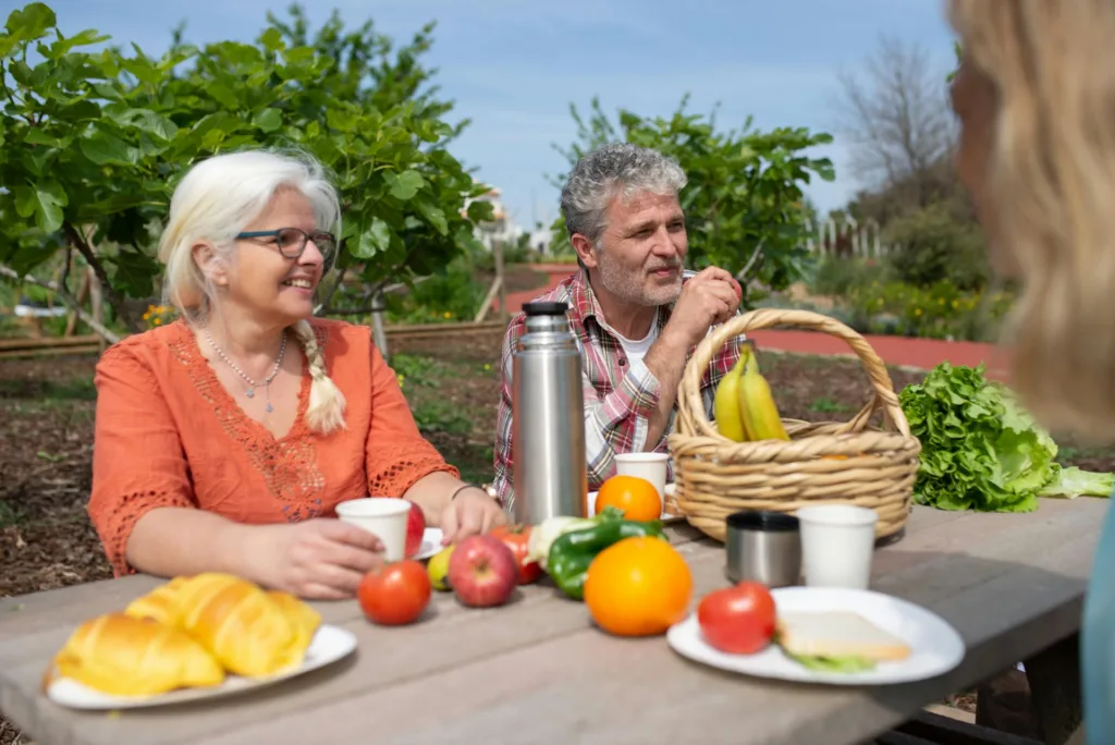 Two elderly people sit on an outdoor picnic bench with food in front of them