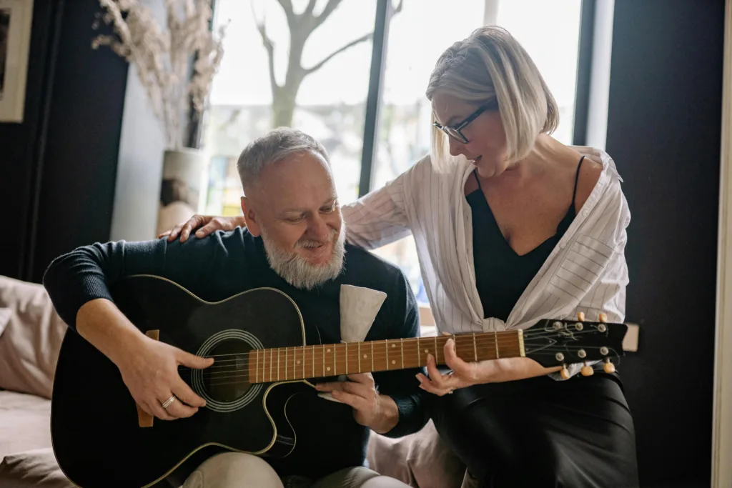 An elderly man plays a guitar on the couch while a lady sits with her arm around him