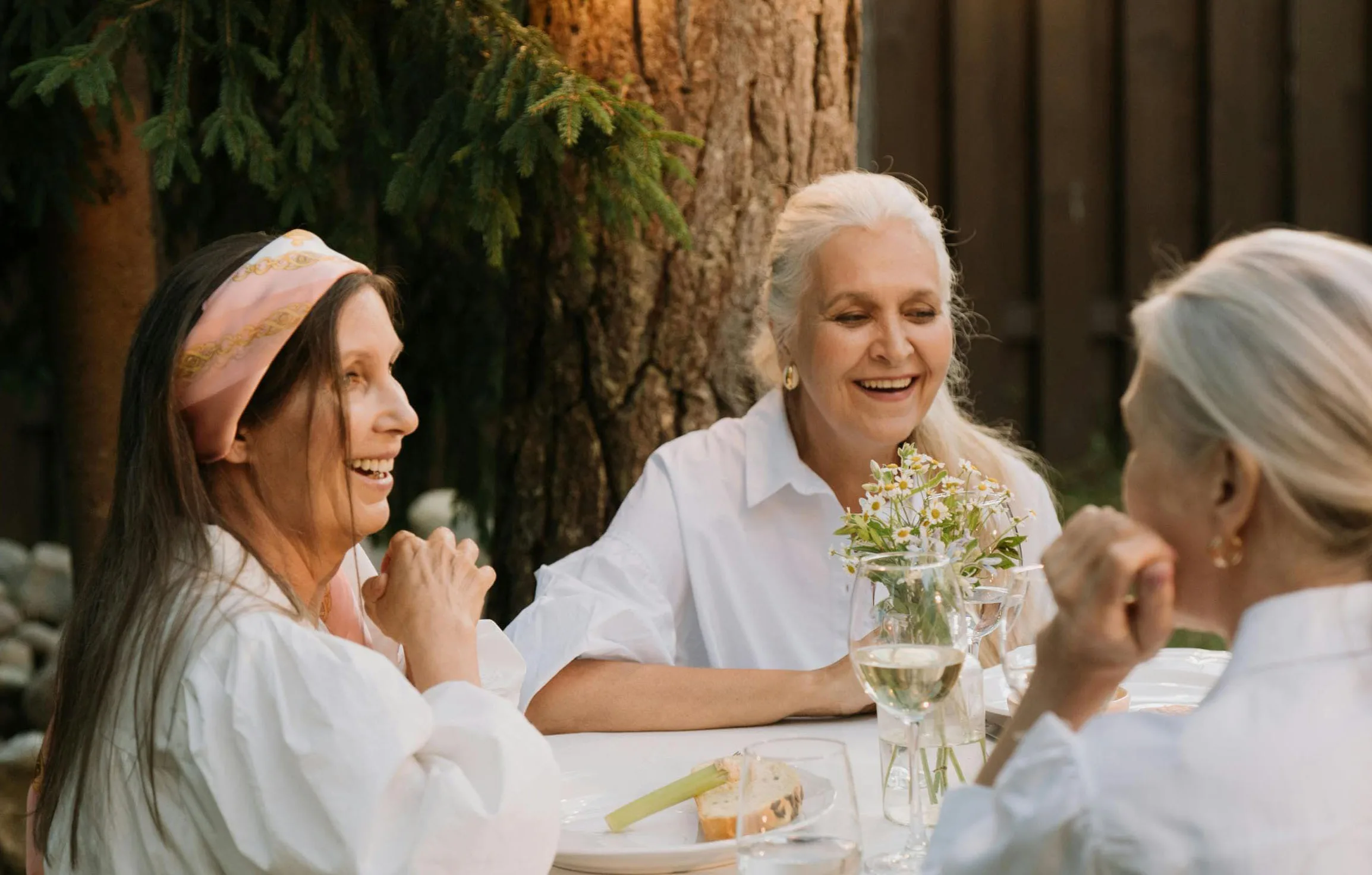 Three smiling women dressed elegantly in white at an outdoor dining table