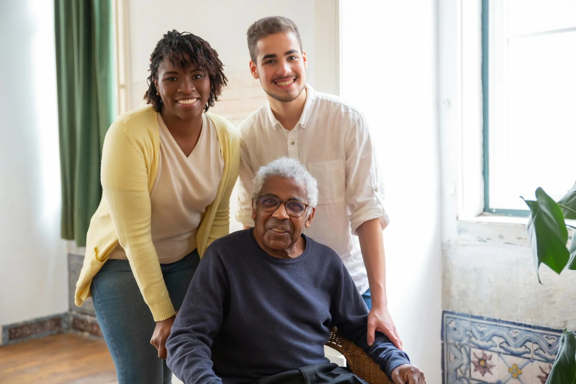 An elderly man sits in a chair with two smiling caretakers behind him